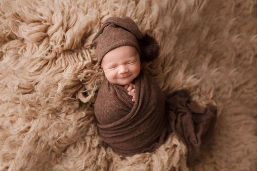 Smiling newborn wrapped in a cozy brown swaddle on a fluffy blanket.